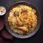 Overhead shot of chicken biryani served on copper plate with yogurt raita on the side.