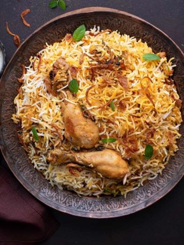 Overhead shot of chicken biryani served on copper plate with yogurt raita on the side.