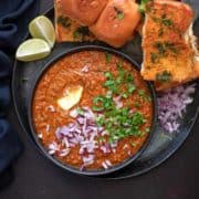 Mumbai Pav bhaji served in black plate with toasted buns, lemon wedges and chopped onions.