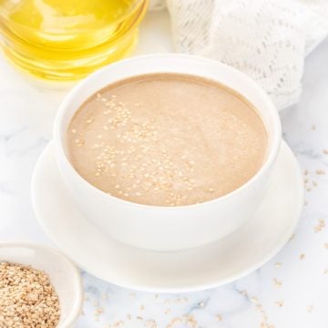 Tahini paste in white bowl, toasted sesame spread around and olive oil at the back.