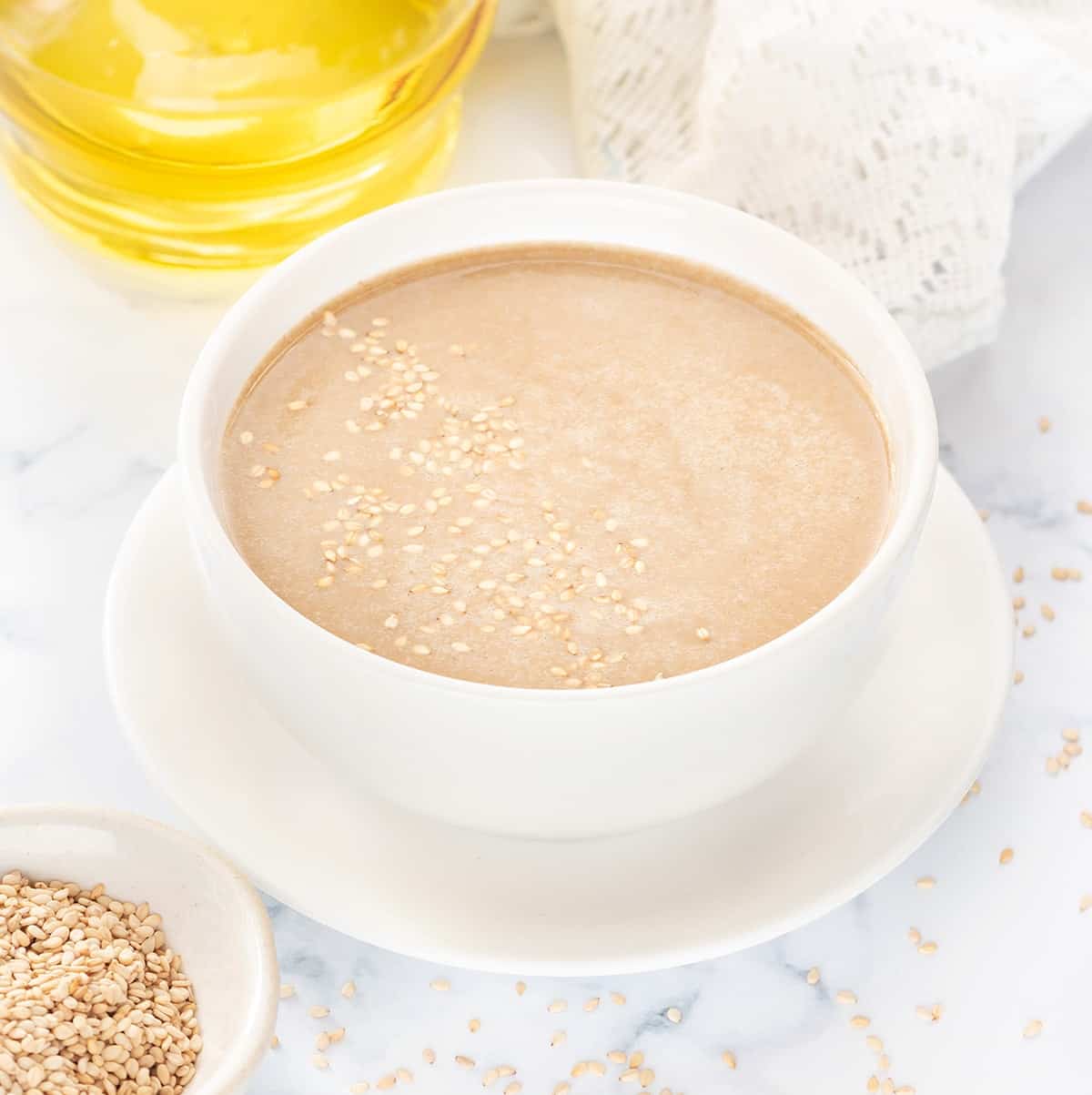 Tahini paste in white bowl, toasted sesame spread around and olive oil at the back.