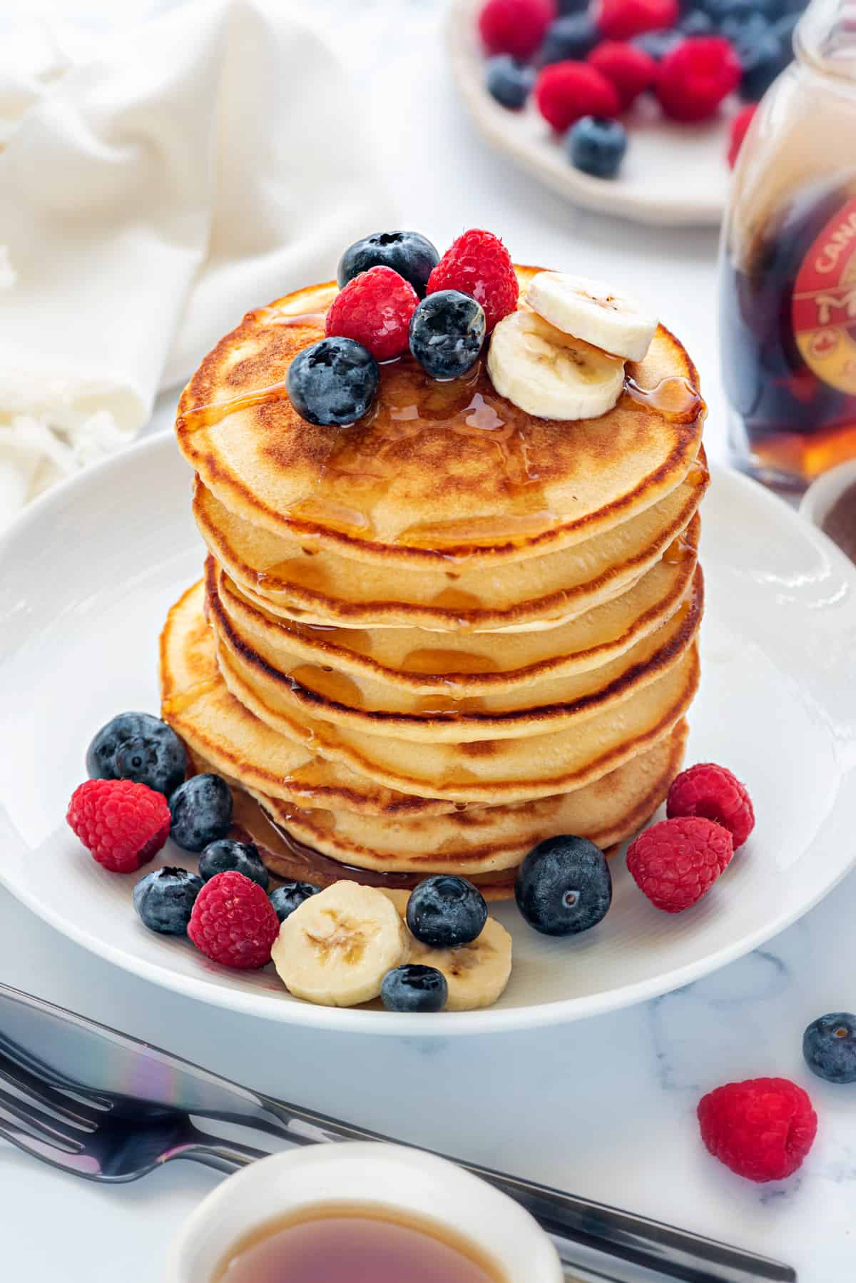 Stacked pancakes topped with maple syrup in white plate with berries, fork and knife on side.