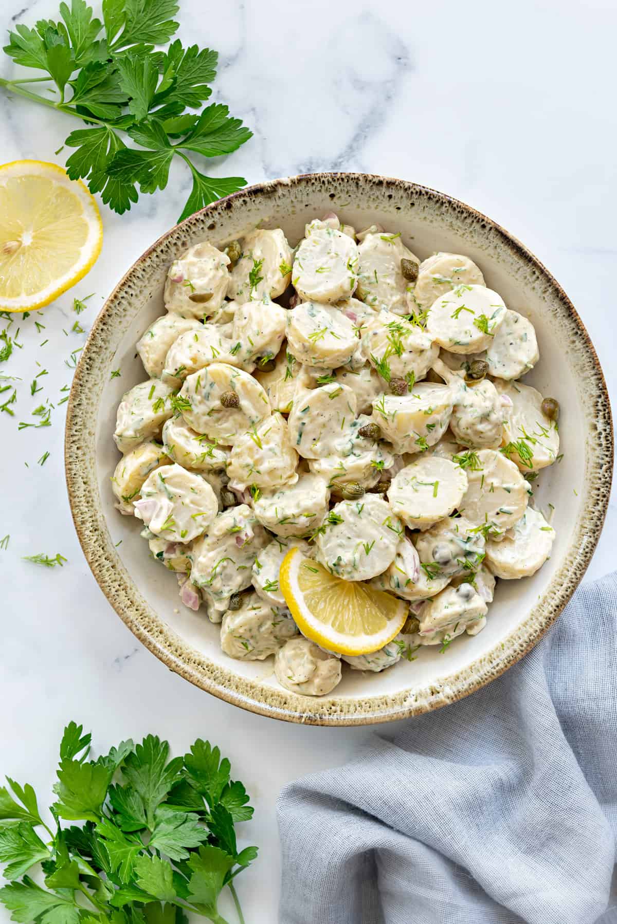 Creamy dill potato salad served in ceramic bowl, lemon slice, dill and parsley spread around.