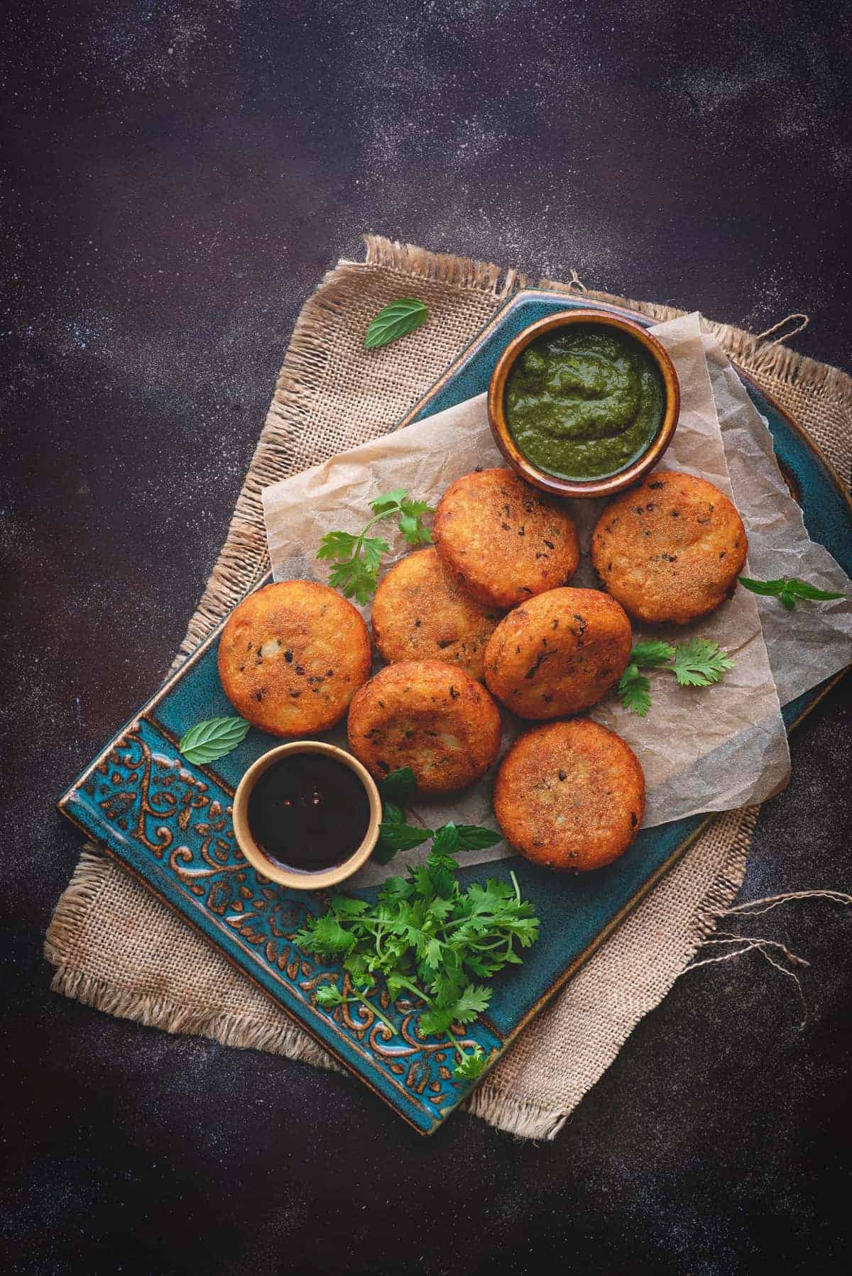 Crispy aloo patties served on  blue ceramic tray, chutney in bowls on side.