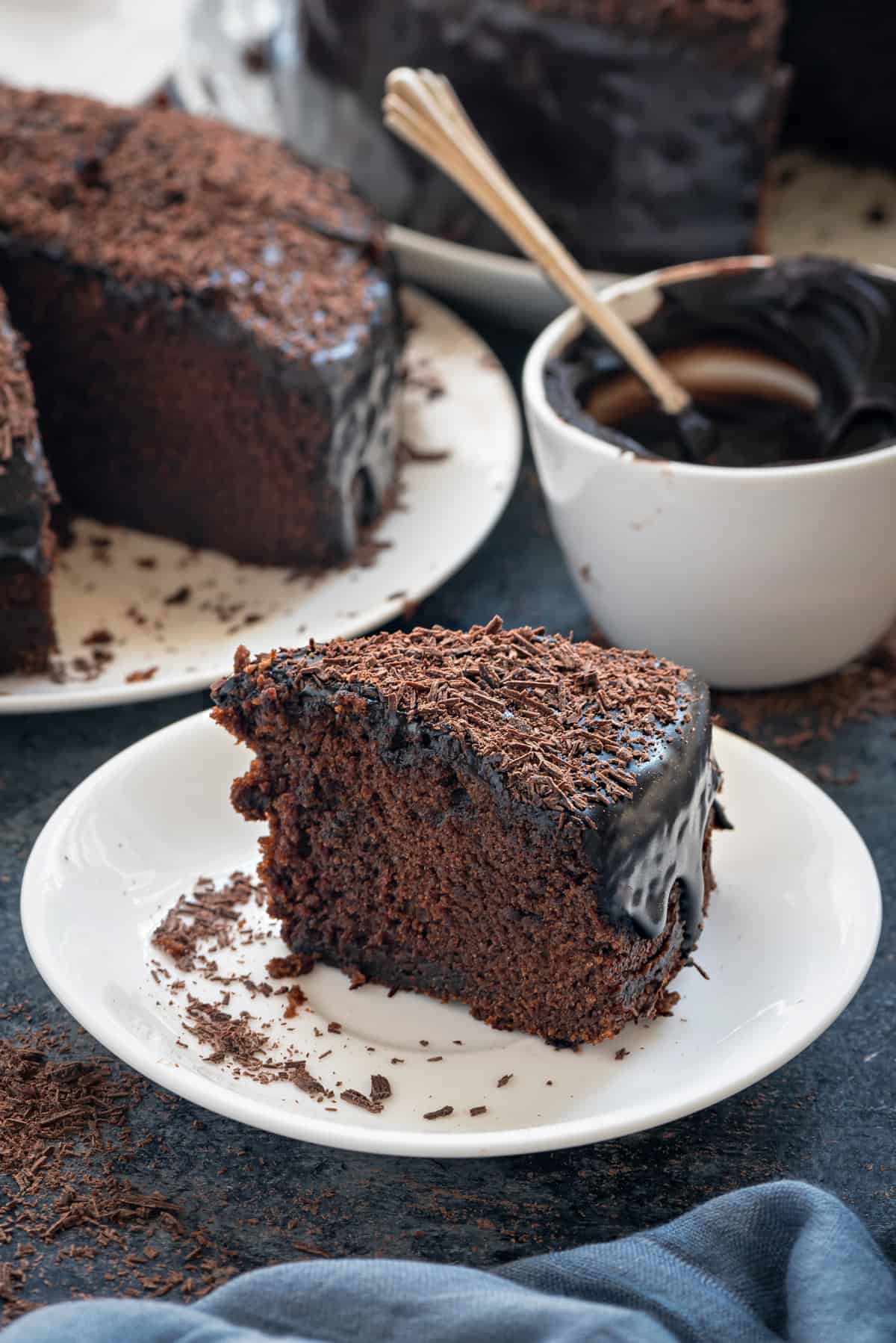 Eggless chocolate cake slice on white plate, white bowl and cake in plate at the back.