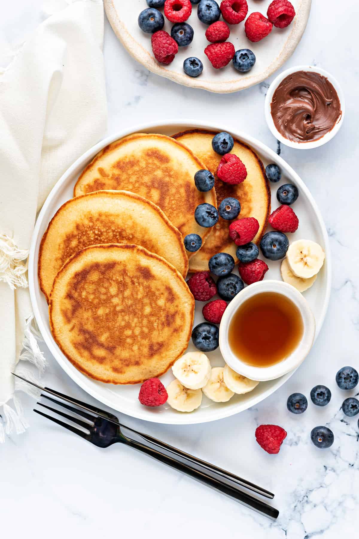 Homemade Pancakes served in white plate with berries banana slices, maple syrup and Nutella on side.