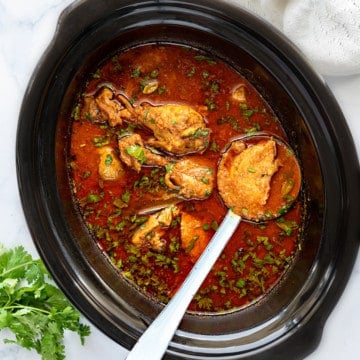 Indian chicken curry in slow cooker pot with a ladle into it holding chicken piece.