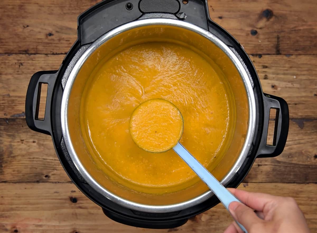 A hand holding a ladleful of curried butternut squash soup in instant pot.