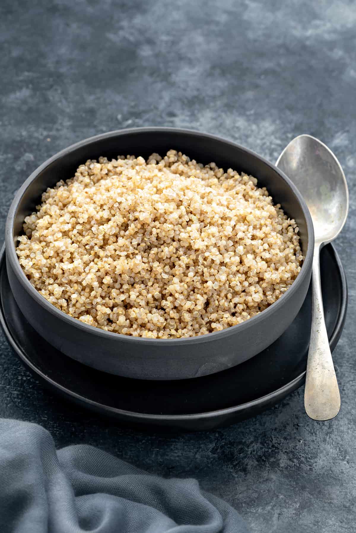 Cooked quinoa served in black bowl with spoon, grey linen on side.