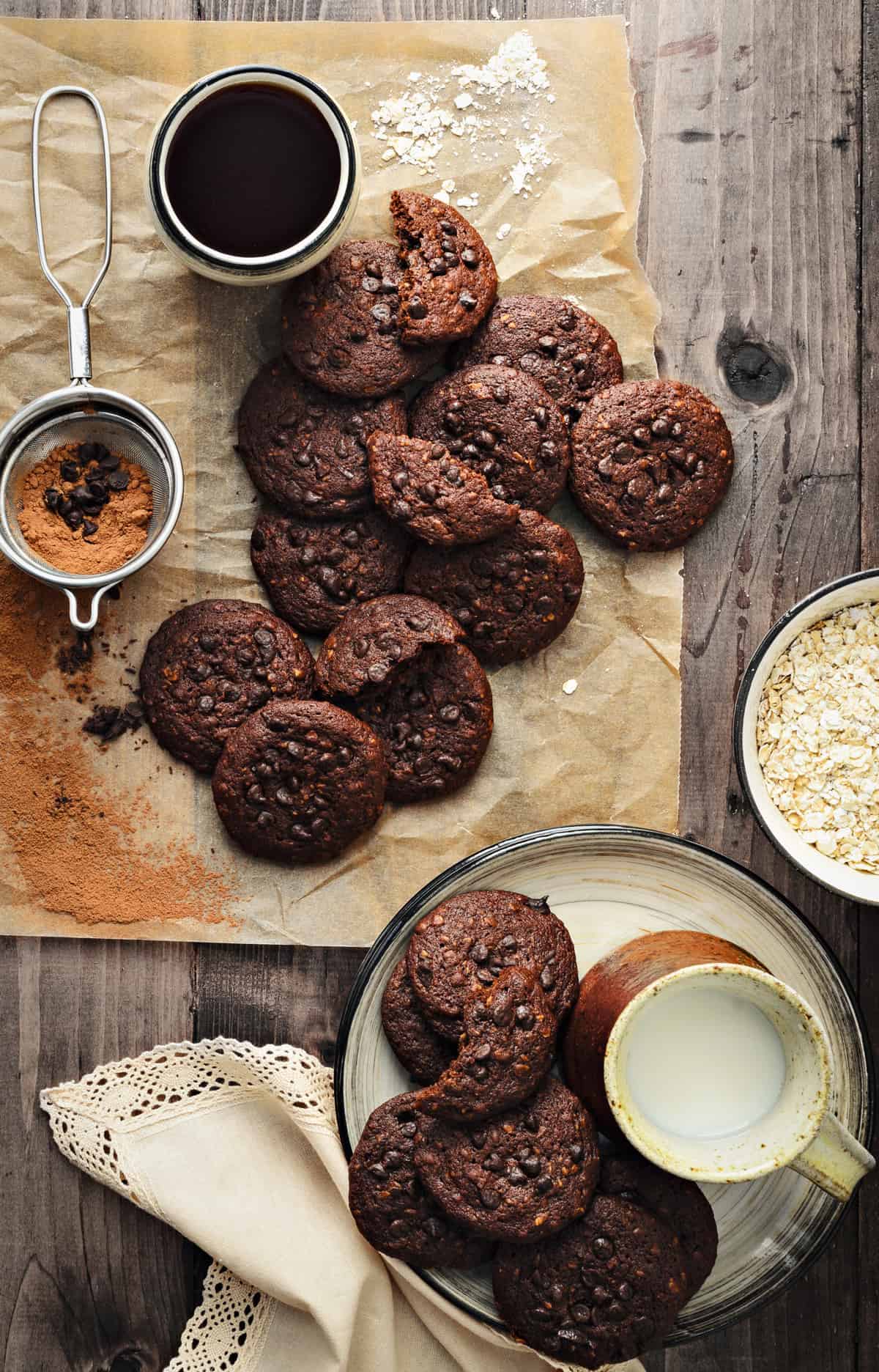 Chocolate Oats cookies on brown paper and plate, with milk, coffee and oats on side.