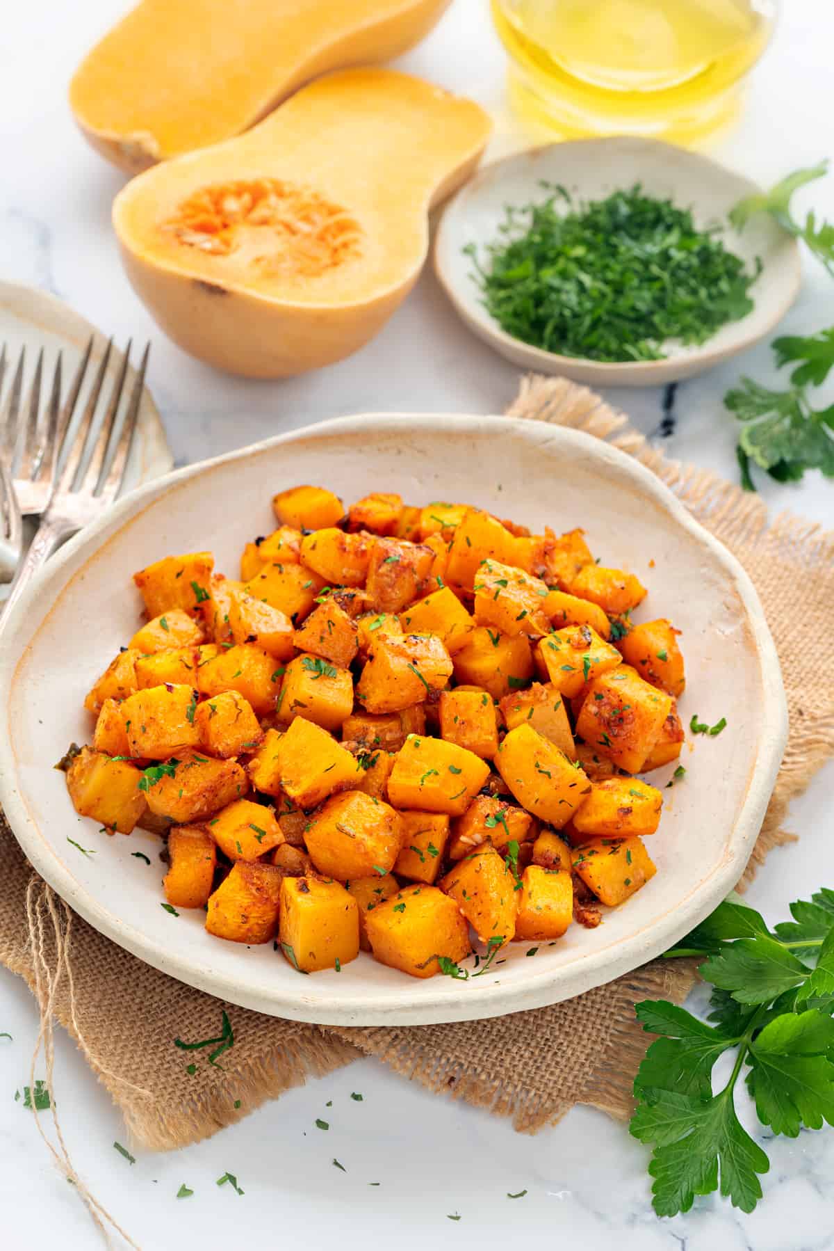 Roasted butternut squash cubes in white ceramic plate, cut squash and parsley placed at the back.