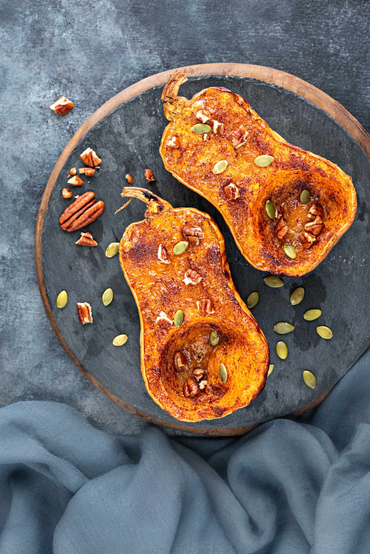 Sweet baked butternut squash halves topped with pecans and pumpkin seeds placed on round black slate.