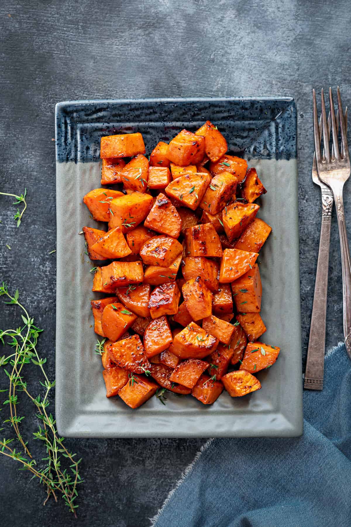 A large plate of roasted sweet potatoes, shot from above, with two forks and sprigs of thyme on the table next to the plate. 