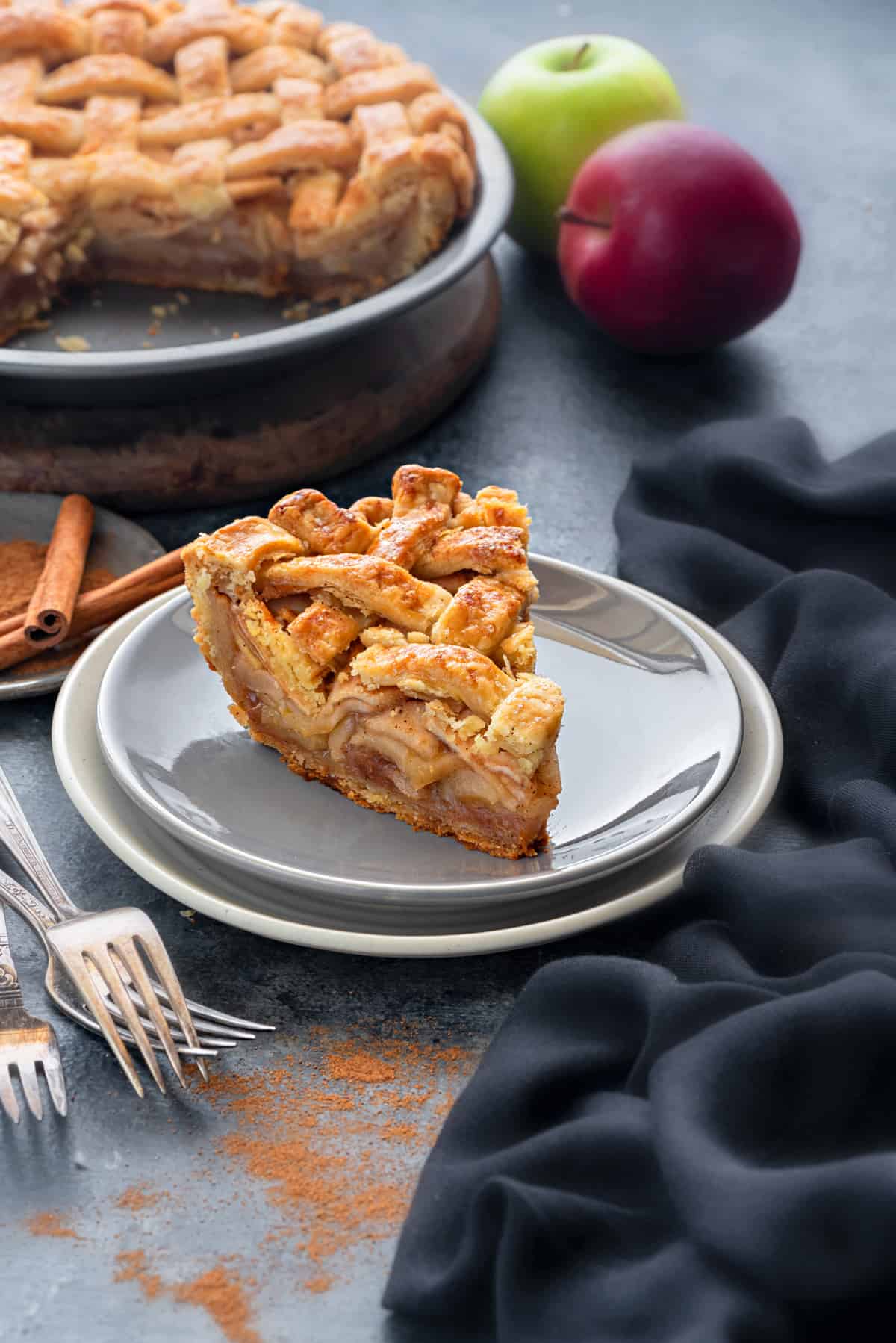 A slice of apple pie on a grey plate, napkins and silverware on the table nearby.