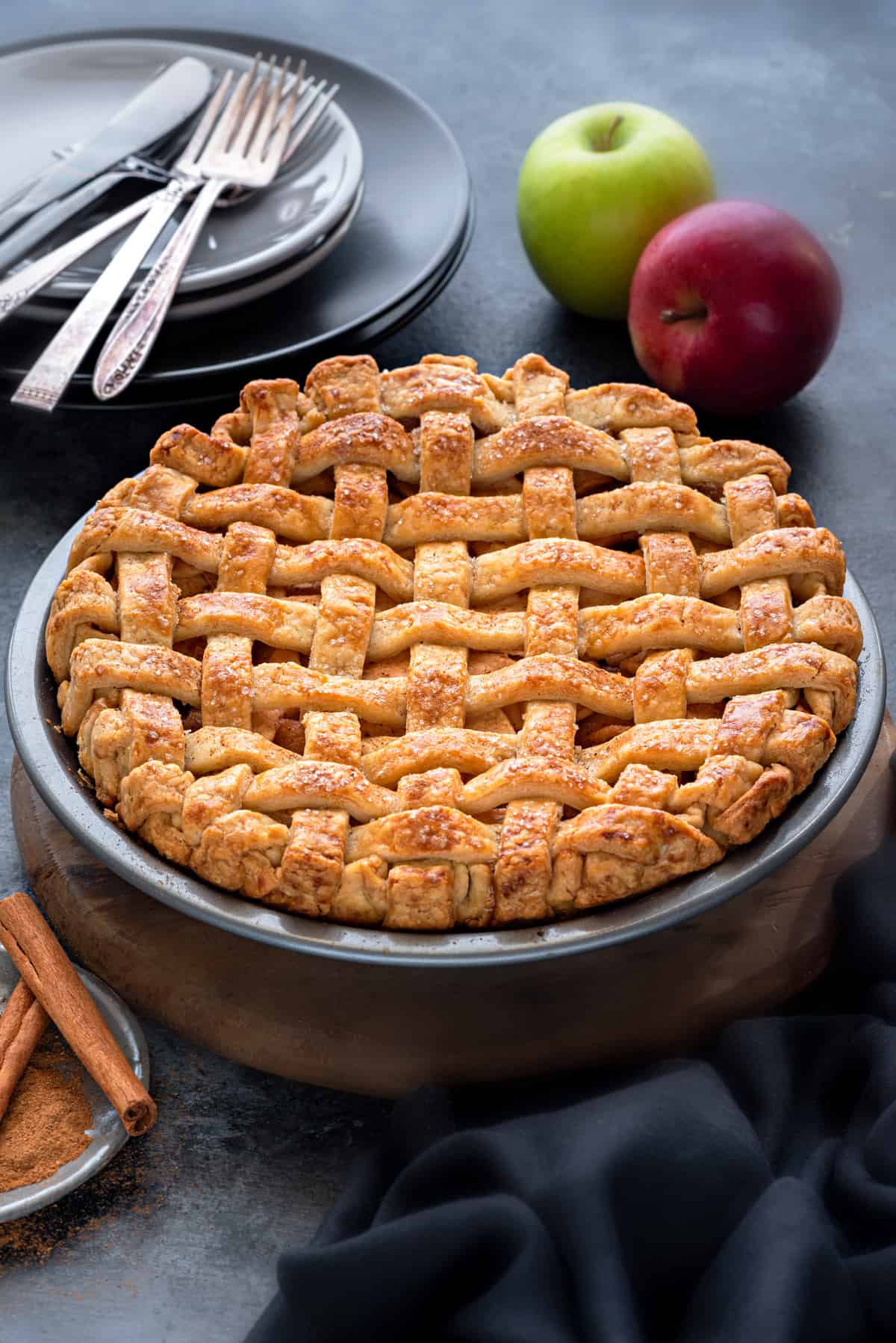 An apple pie with crispy lattice crust, shot from slightly above, apples placed in the background.