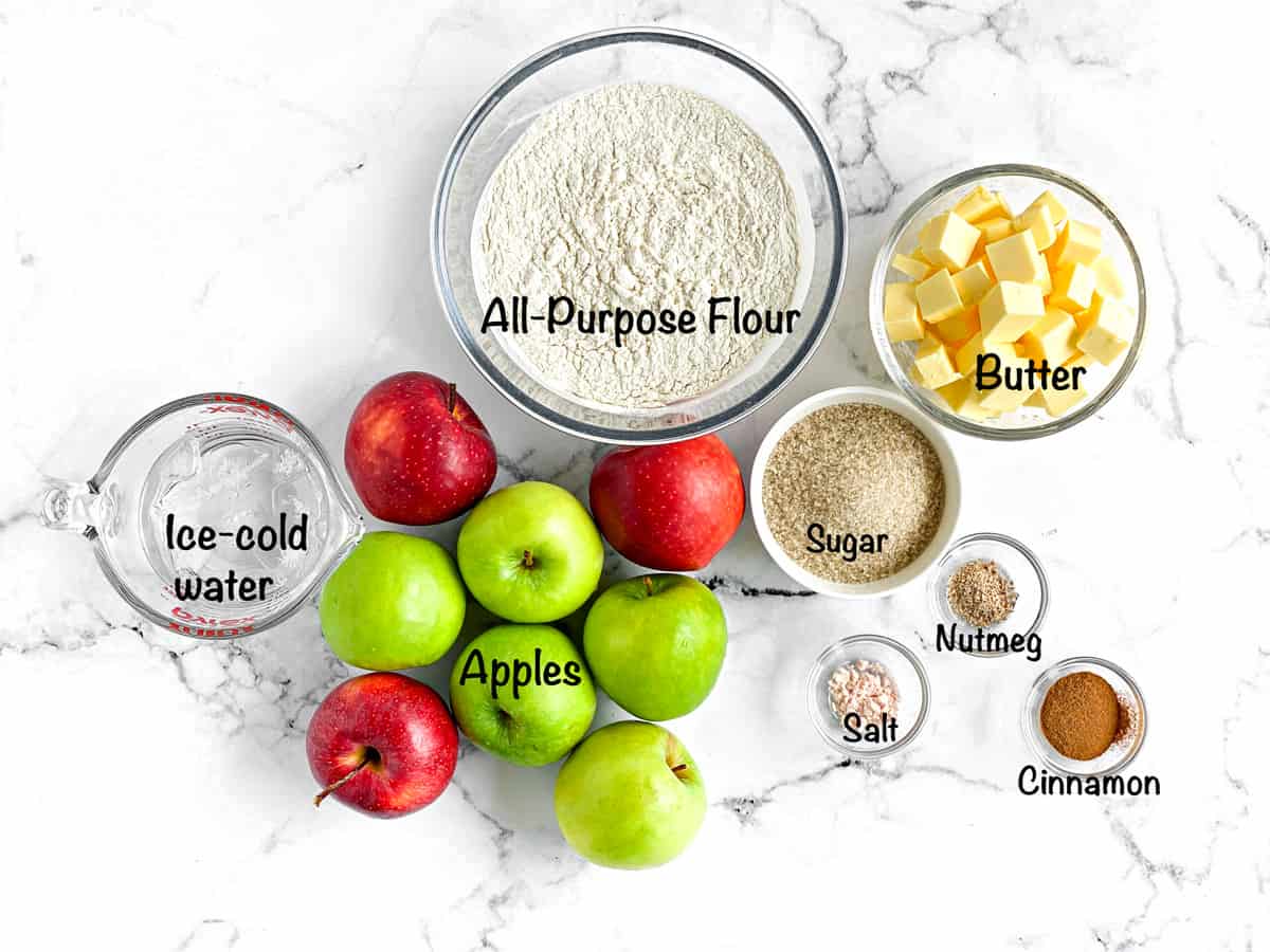 Overhead shot of the ingredients for this pie. Water, apples, flour, butter, sugar, nutmeg, salt, cinnamon. 