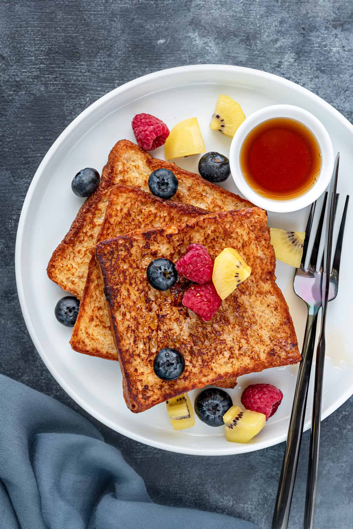 Classic brioche french toast on a round white plate with silverware, maple syrup and fresh fruits.