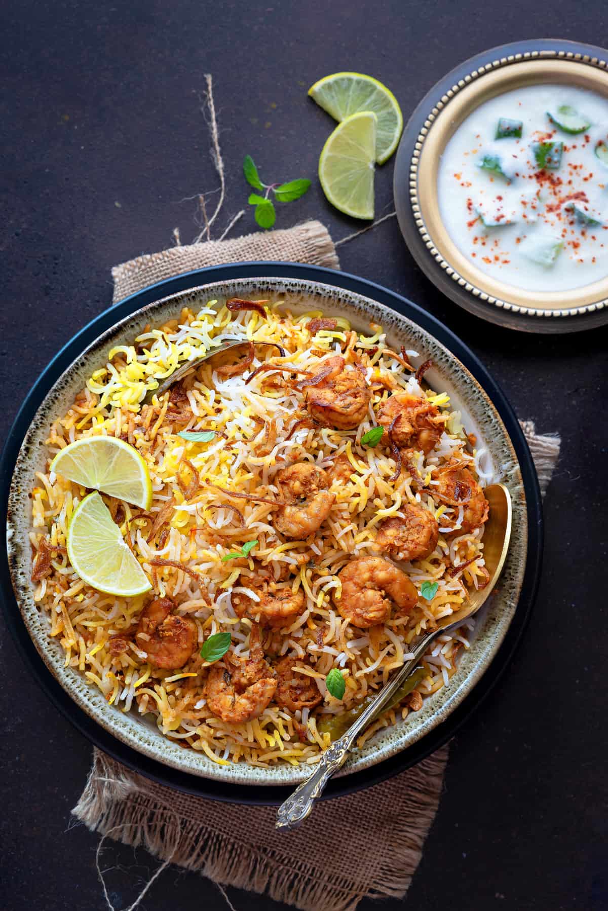 Overhead shot of prawn biryani in bowl with a spoon and yogurt raita on the side.