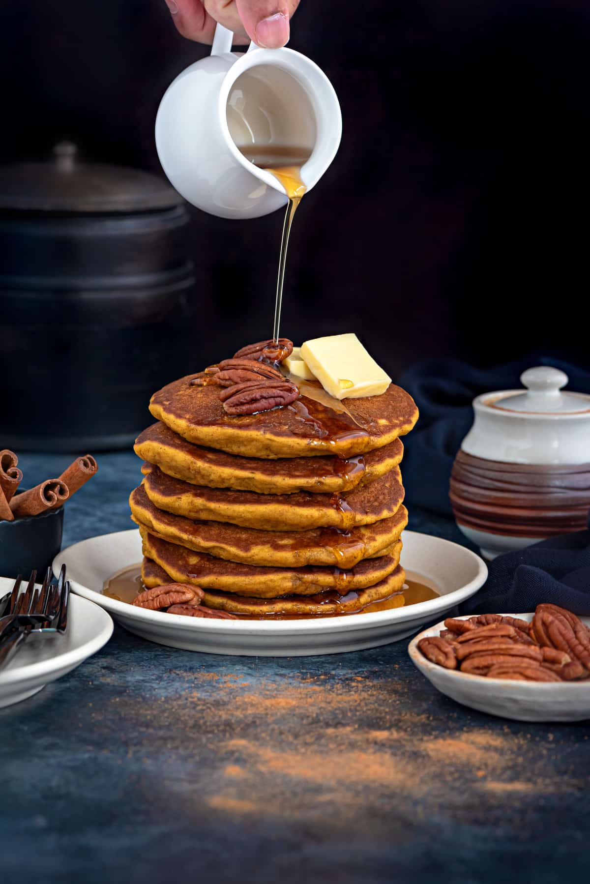 stack of pumpkin pancakes on a white plate with a hand pouring maple syrup over them