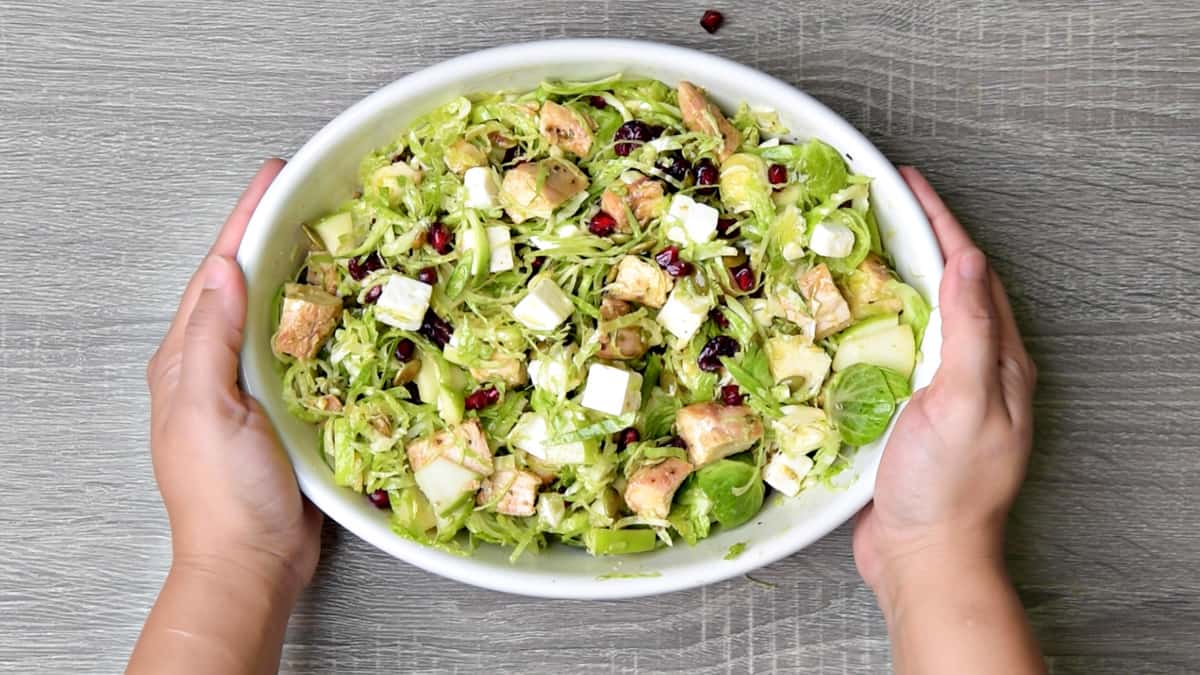 two hands holding white serving bowl with shaved brussels sprouts salad