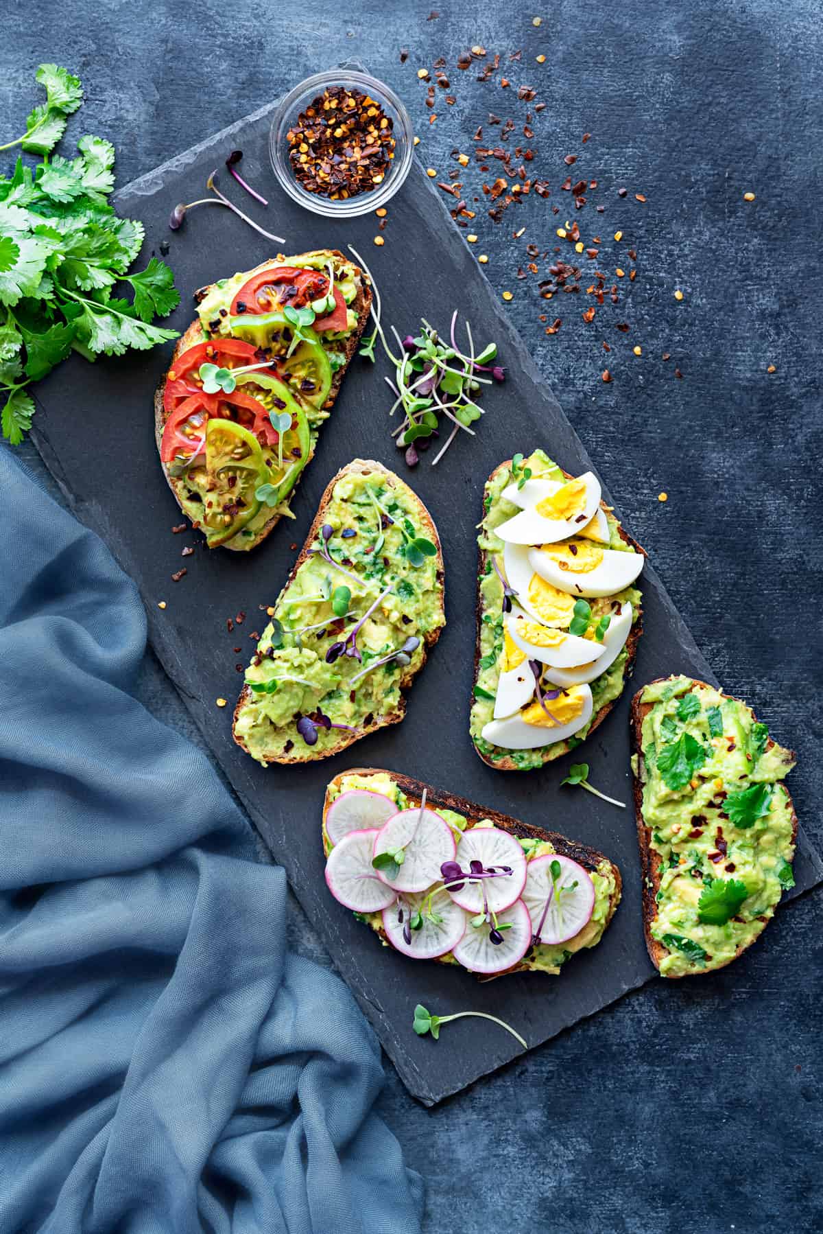 overhead shot of avocado toast with different topping ideas - microgreens, boiled egg, sliced radishes, and tomatoes