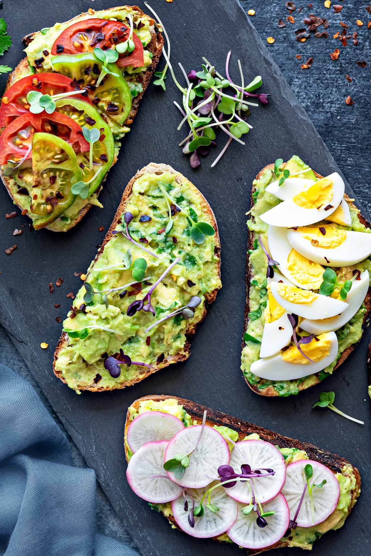 overhead shot of four slices of avocado toast with different toppings on a black slate serving tray