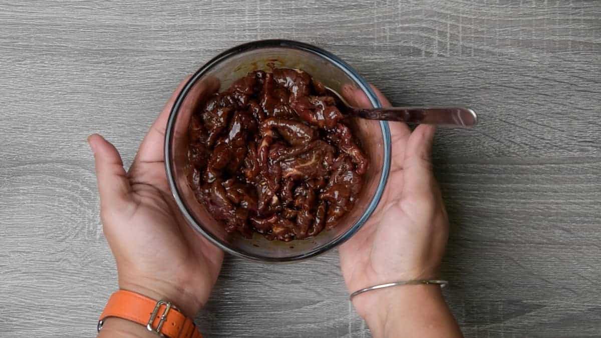 two hands around clear glass bowl with beef tossed in stir fry marinade