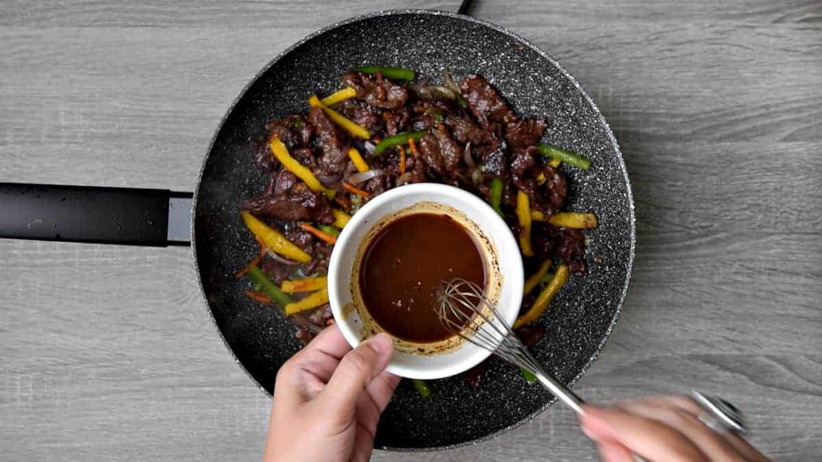 hand holding whisk with bowl of rice vinegar, water and extra marinade above stir fry in wok