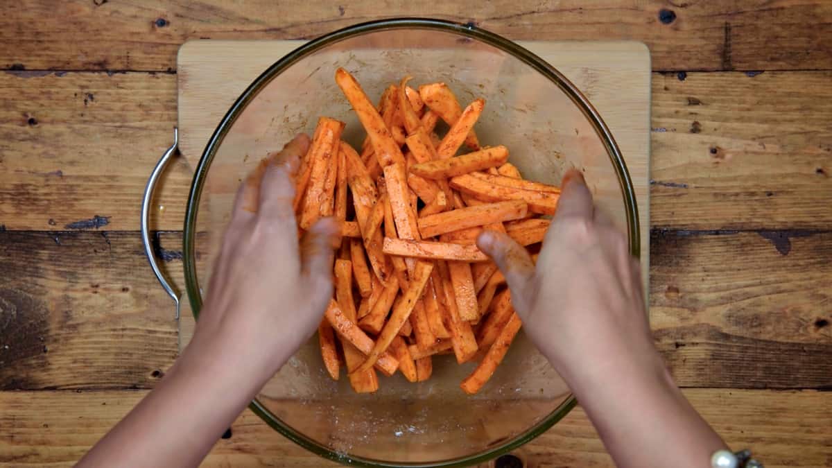 hands mixing together sweet potato batons with oil and seasonings in a glass mixing bowl