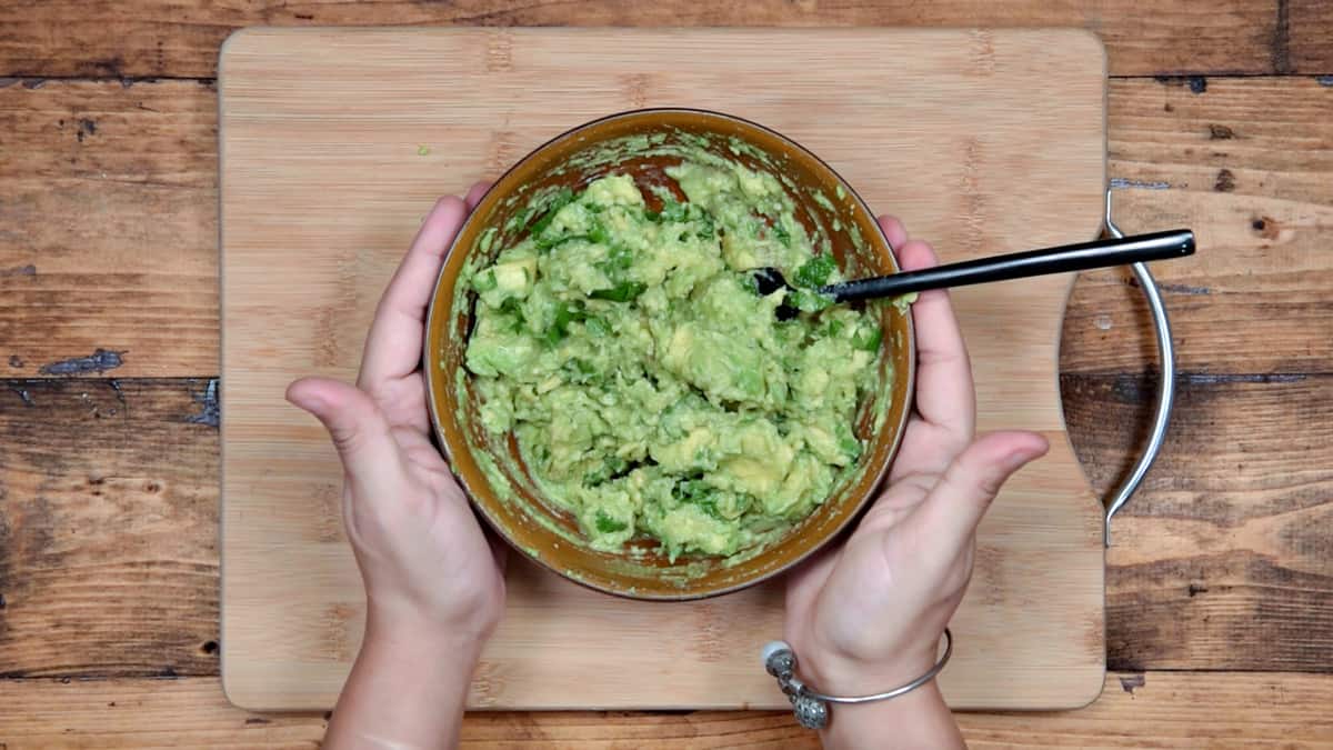 two hands holding a brown bowl of avocado mashed with cilantro and lime 