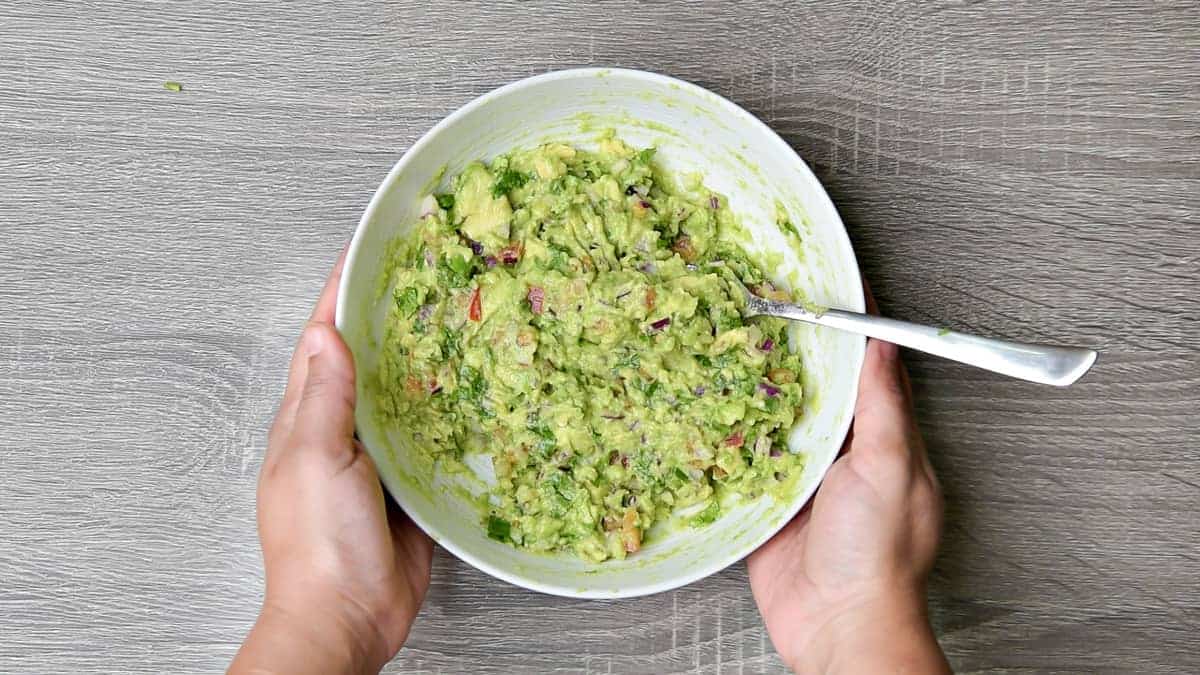 hands holding white bowl with completed recipe for guacamole recipe