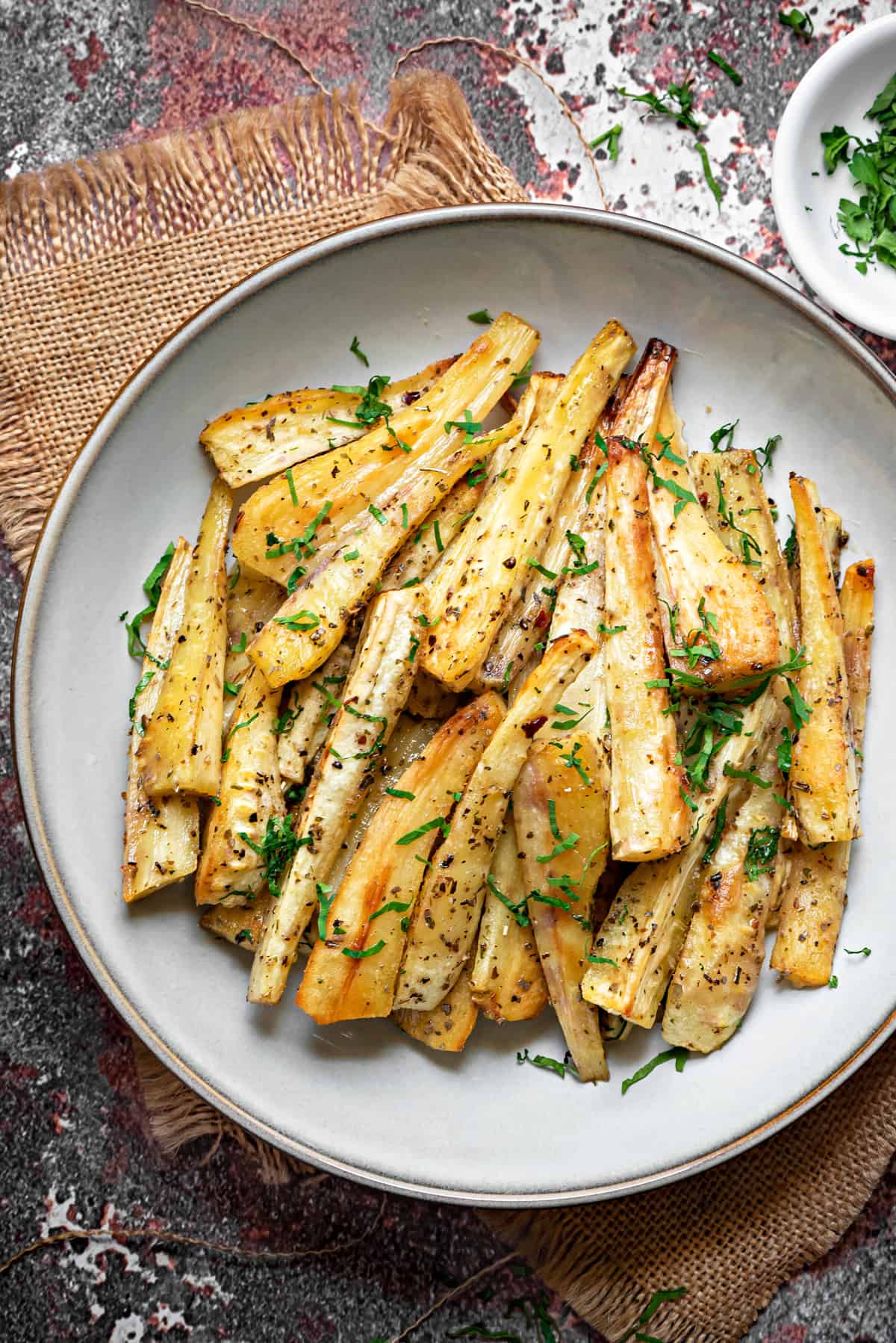 overhead shot of a pile of roasted parsnip batons on a white plate and sprinkled with fresh parsley