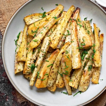 A pile of roasted parsnip batons on a white plate sprinkled with fresh parsley