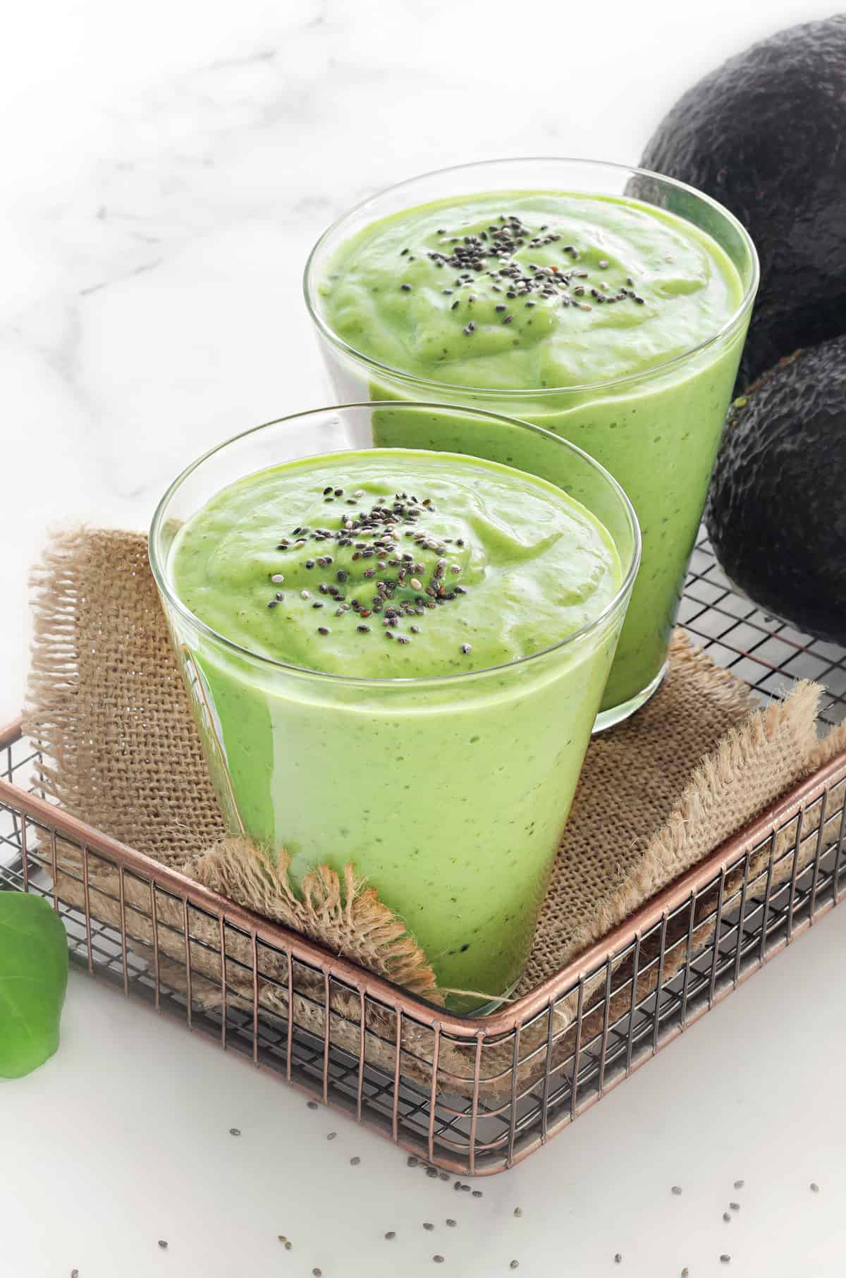 two glasses of healthy green smoothies topped with chia seeds with avocados in the background