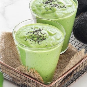 two glasses of healthy green smoothies topped with chia seeds with avocados in the background