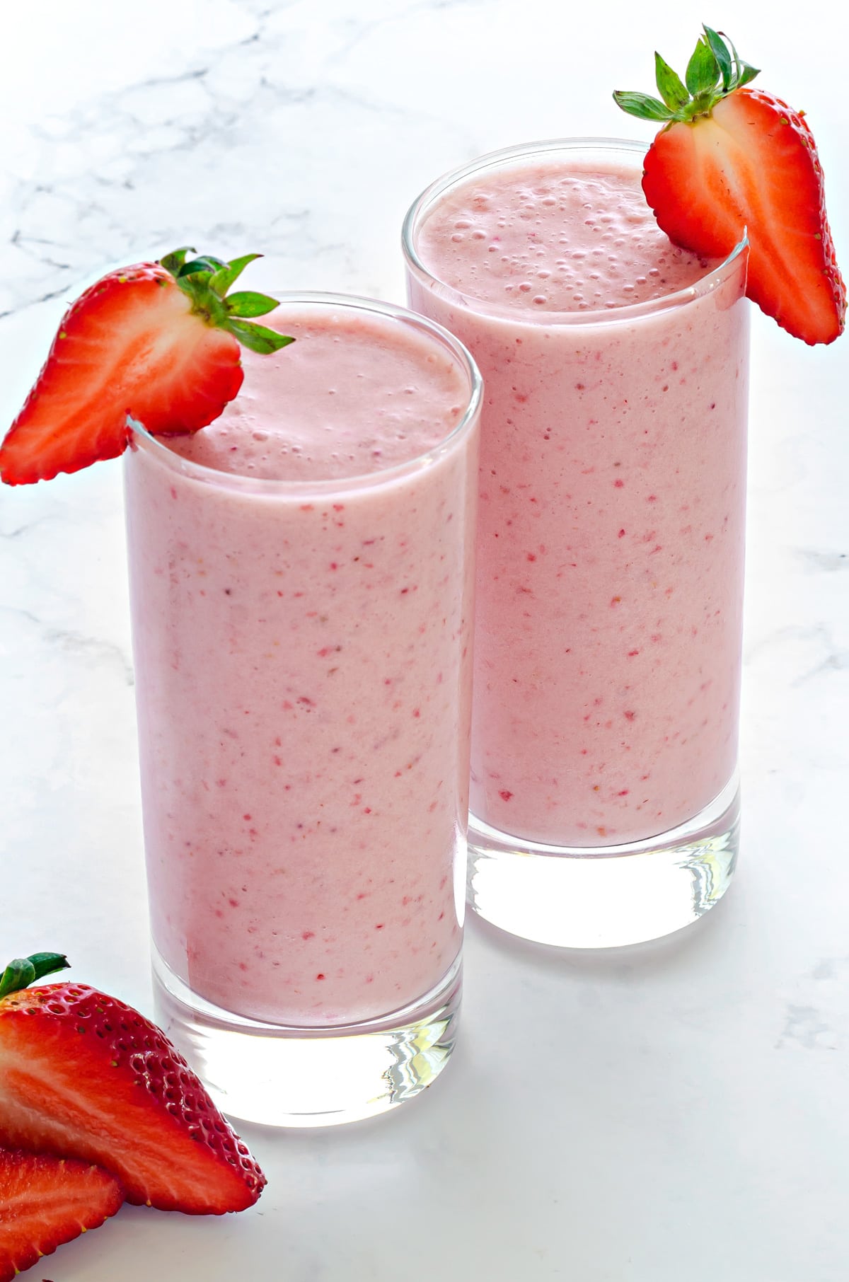 two tall clear collins glasses filled with strawberry smoothie and garnished with fresh strawberry slices