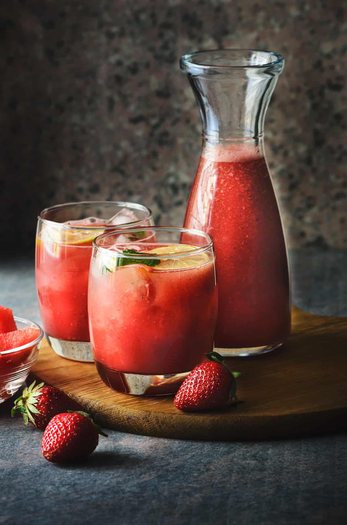 glass carafe and two rocks glasses filled with watermelon strawberry lemonade and fresh strawberries scattered around