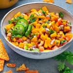 mango salsa in a white earthenware bowl next to a plate of tortilla chips and mangoes
