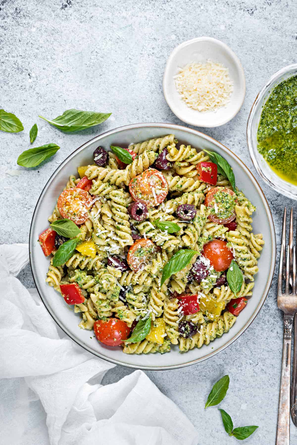 A serving bowl filled with pesto pasta salad and silver forks on a grey table