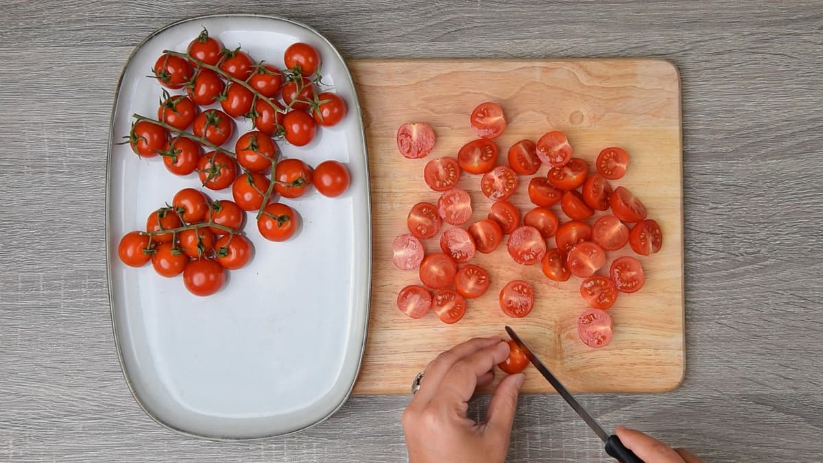 hands slicing roughly half of the cherry tomatoes on a wooden cutting board