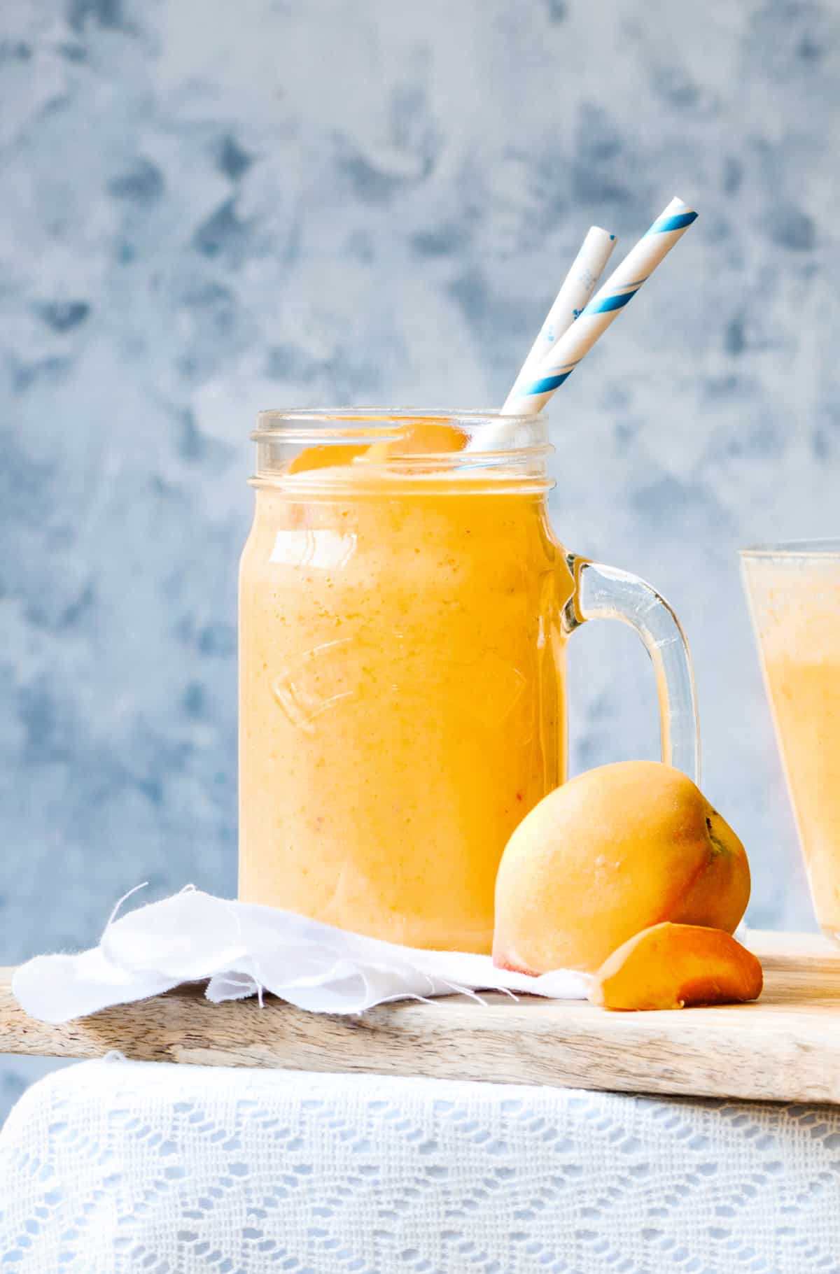 glass pitcher filled with peach smoothie on a wooden board with a fresh peach in the foreground