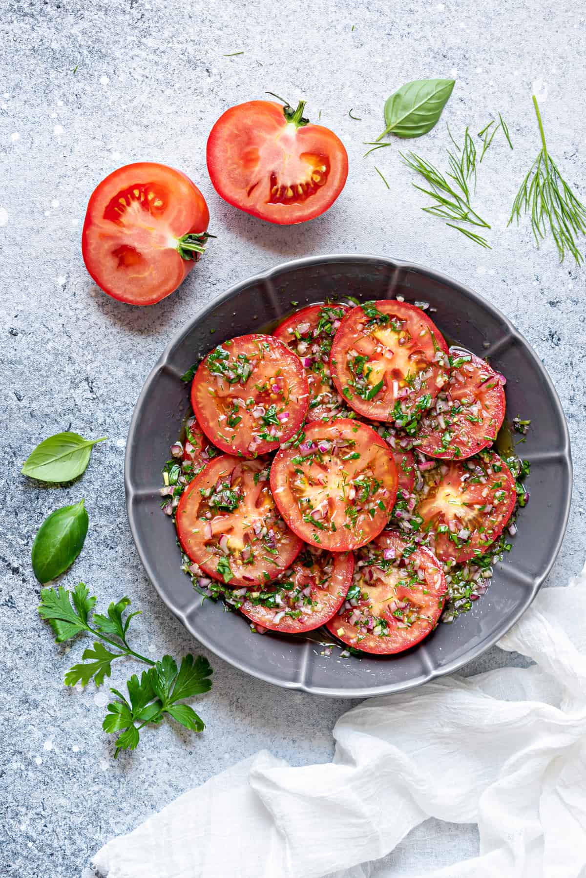 marinated tomatoes in grey serving plate with some fresh herbs spread around on a white table