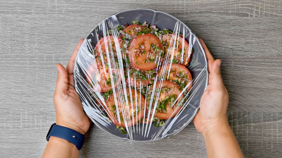 two hands holding plastic wrapped grey dish filled with marinated tomatoes 