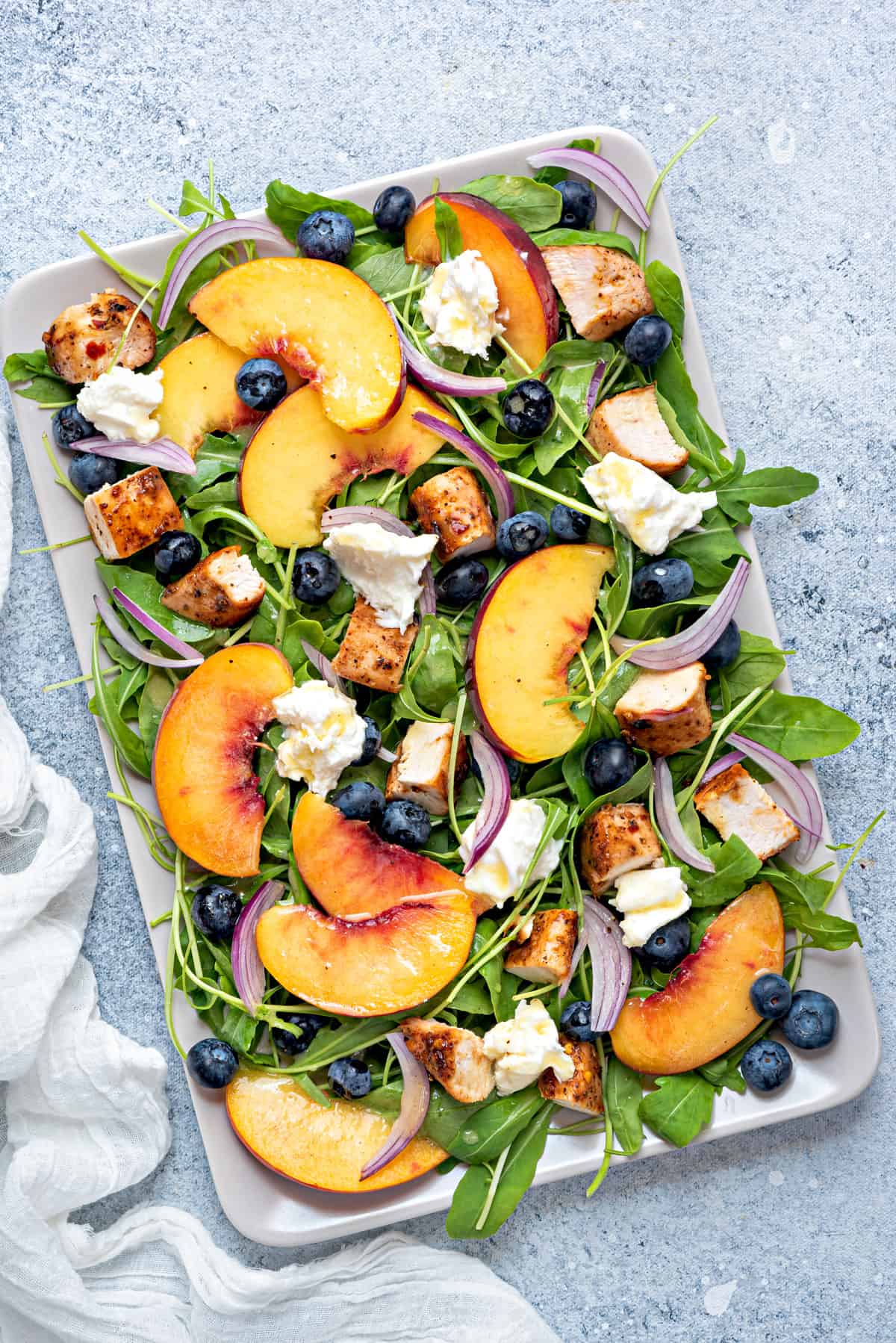 a rectangular serving platter of peach arugula and burrata salad with red onions, chicken and blueberries
