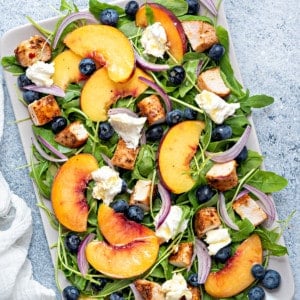 a rectangular serving platter of peach arugula and burrata salad with red onions, chicken and blueberries