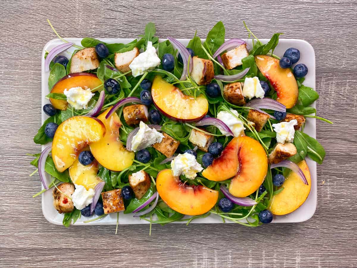 overhead shot of peach arugula salad after drizzling on the dressing and adding the burrata, red onion and berries