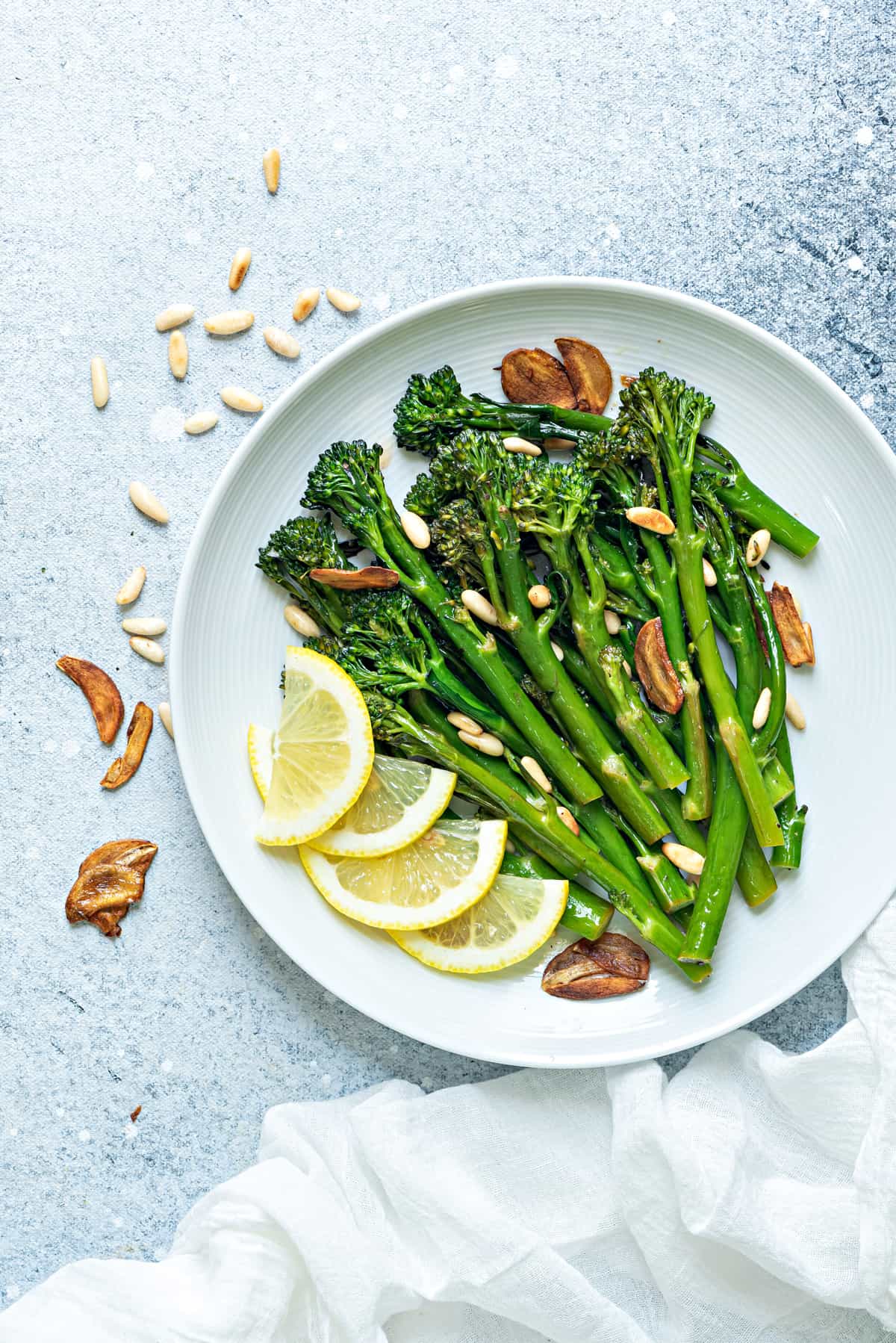 a serving platter of sautéed broccolini with fresh lemon slices, garlic chips and toasted pine nuts