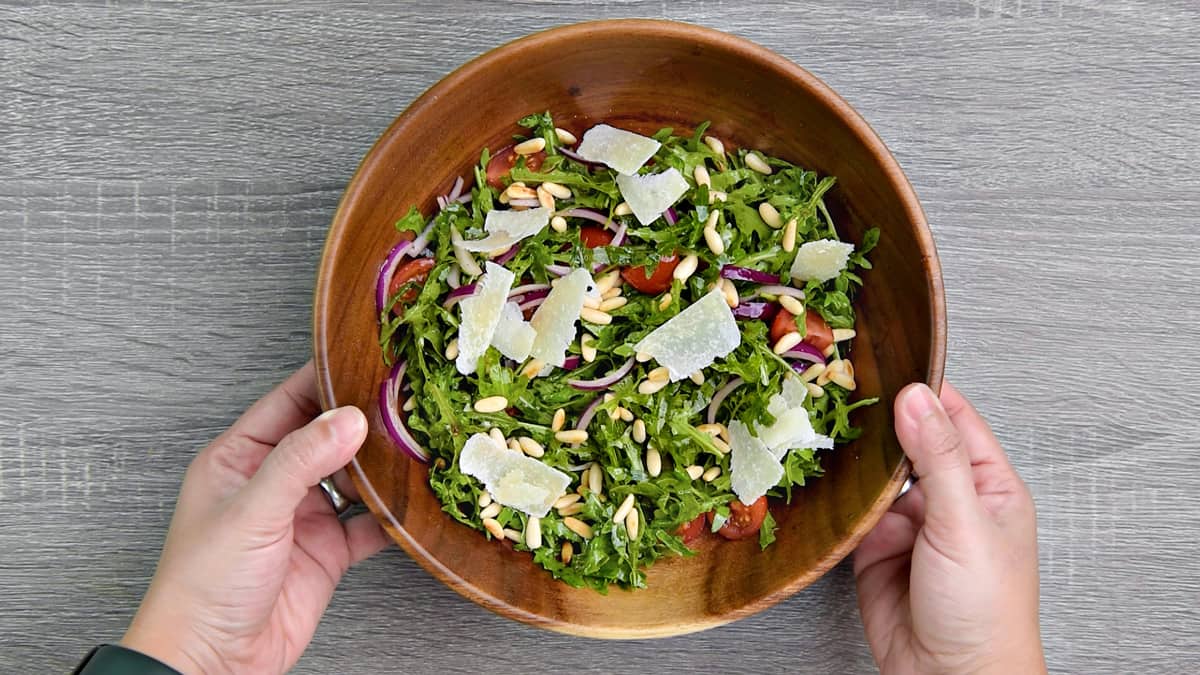 hands holding a wooden bowl with arugula salad servings garnished with pine nuts and parmesan shavings