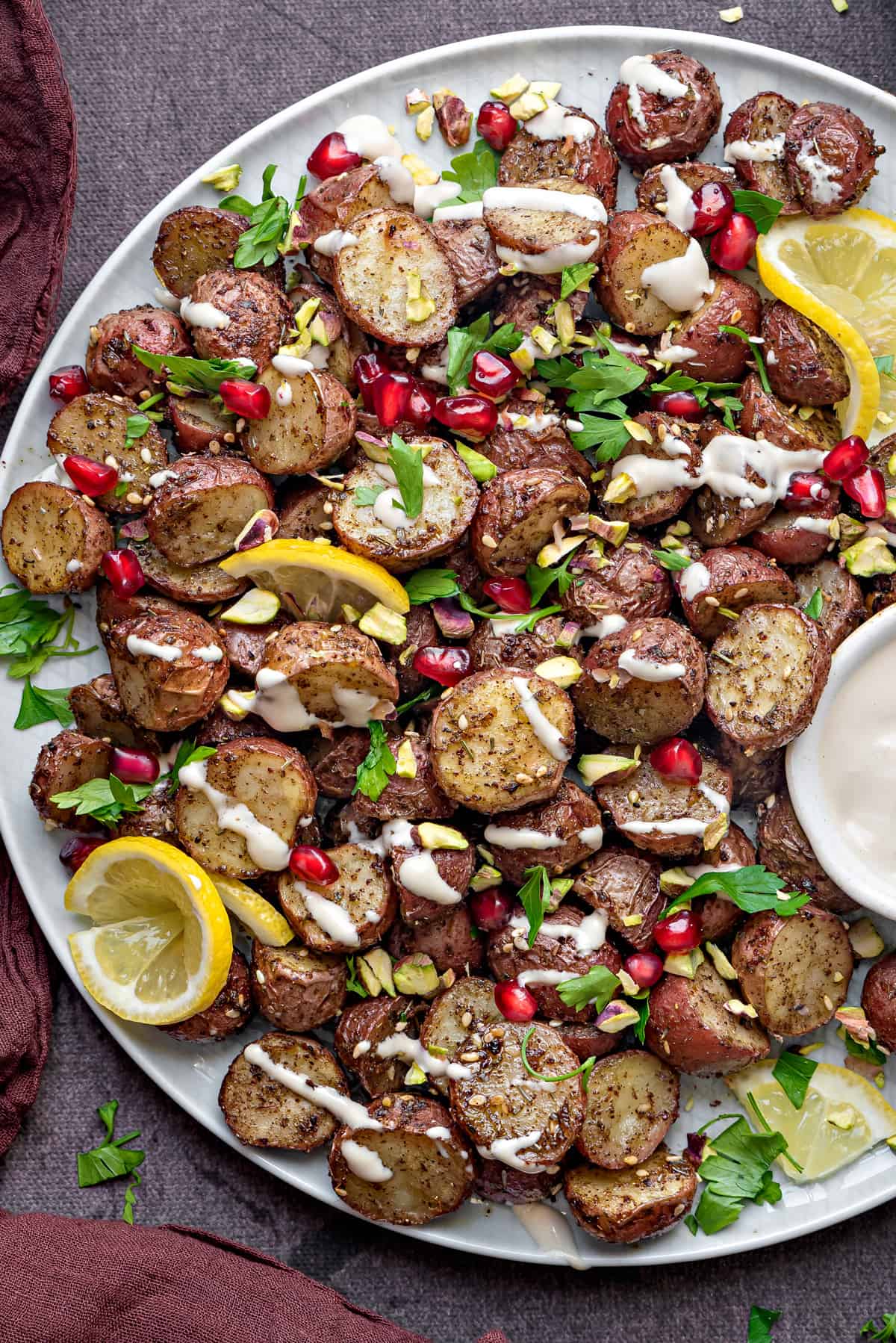 white plate with roasted red potatoes, garnished with pomegranate, tahini sauce, lemon, parsley, pistachios