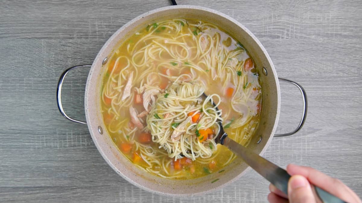 hand holding a ladle with chicken noodle soup in pot.